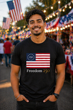 Man wearing a black t-shirt with a flag design and 'Freedom 250' text, standing in front of festive lights and American flags.