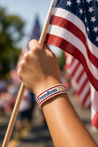 Hand holding an American flag with a 'Freedom 250' wristband, blurred background