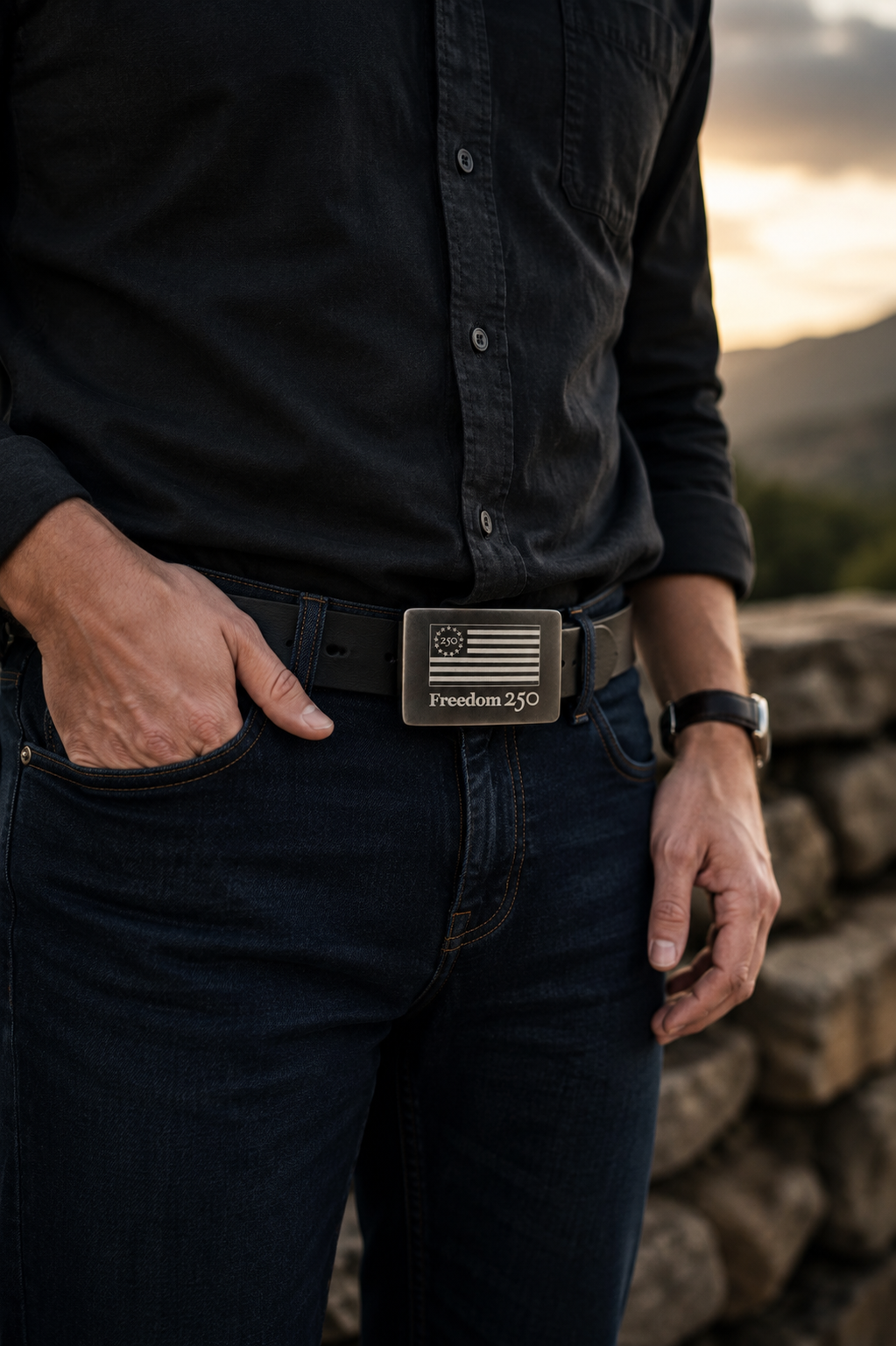Person wearing a black shirt and jeans with a belt featuring a visible brand logo, standing against a sunset background.