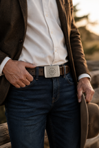 Person wearing a brown jacket, white shirt, blue jeans, and a belt with a decorative buckle.