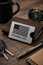 Metal card with American flag design and 'Freedom 250' text on a wooden surface with wallet, keys, pen, and watch.