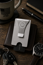 Metal money clip on a wooden surface with a mug, pen, wallet, keys, and watch.