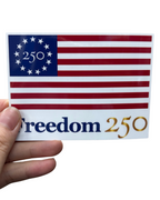 Sticker with American flag design and 'Freedom 250' text held by a hand