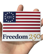 Sticker with American flag design and 'Freedom 250' text held between fingers