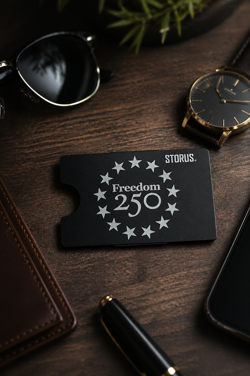 Black card with 'Freedom 250' design on a wooden surface with sunglasses, watch, and phone.